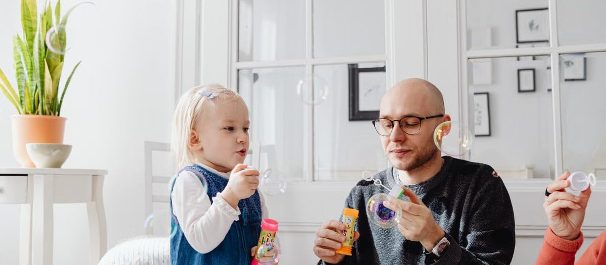 A joyful moment between a father and daughter blowing bubbles indoors, highlighting family fun.