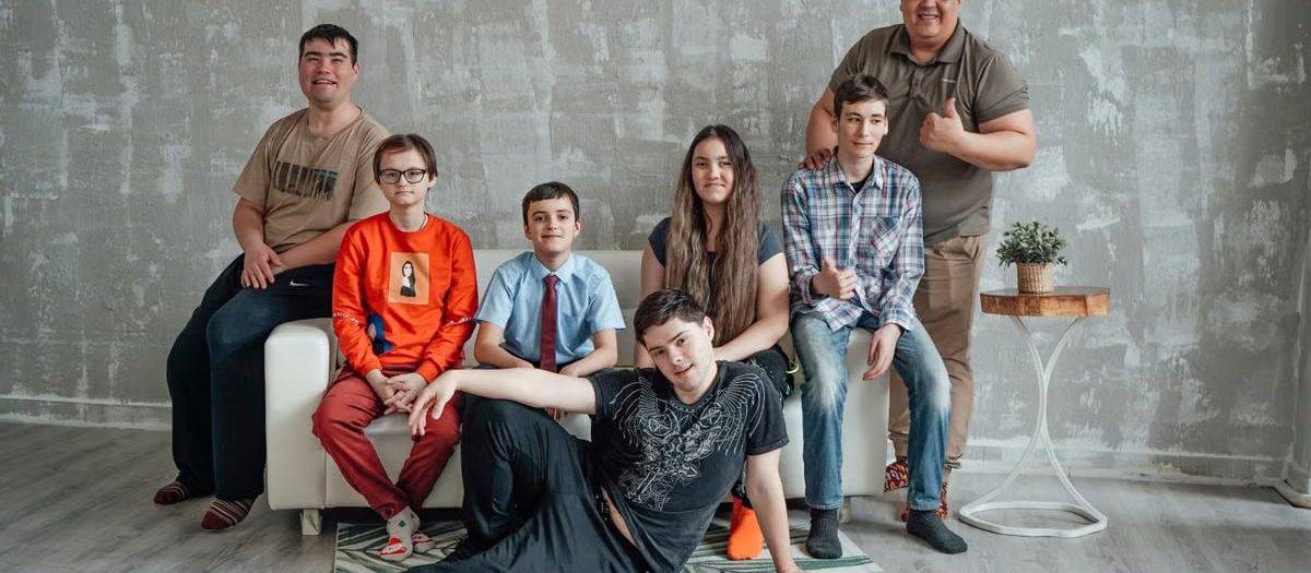 Group of seven people posing casually in a stylish indoor setting with a concrete wall background.