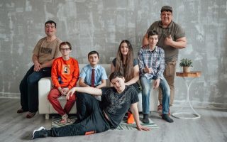 Group of seven people posing casually in a stylish indoor setting with a concrete wall background.